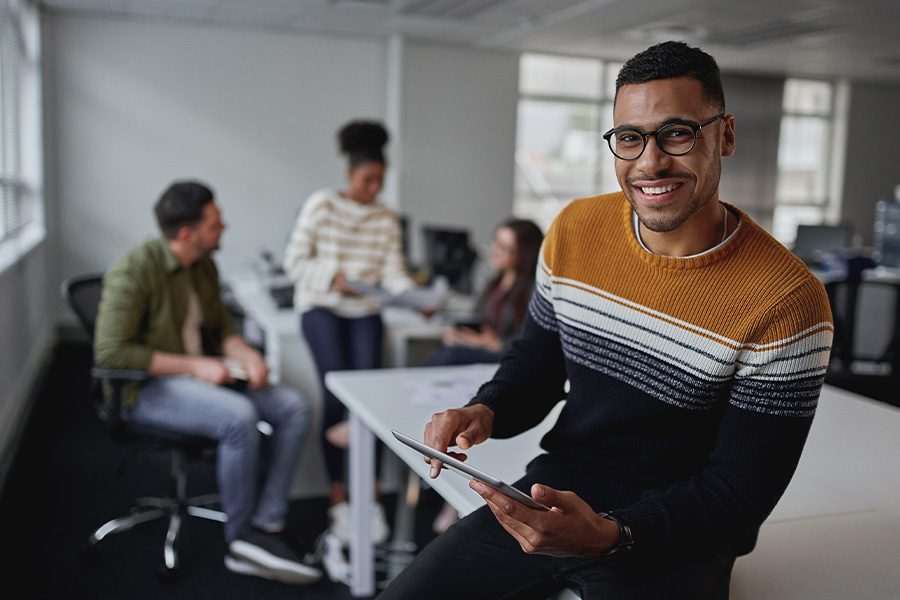 Business Insurance - Portrait of a Successful Smiling Businessman Holding a Tablet Looking at Camera and Sitting in Front of His Team in a Modern Office